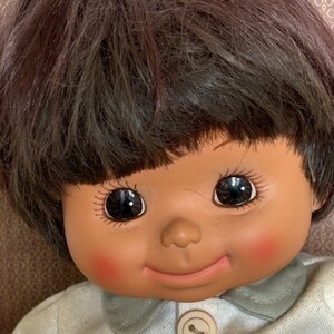 Puede incluir: Una mu&ntilde;eca sonriente con cabello casta&ntilde;o y ojos marrones que lleva una camisa azul claro con dos botones blancos.