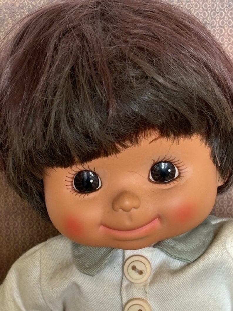 Puede incluir: Una mu&ntilde;eca sonriente con cabello casta&ntilde;o y ojos marrones que lleva una camisa azul claro con dos botones blancos.