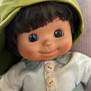 Puede incluir: Una mu&ntilde;eca sonriente con cabello casta&ntilde;o que lleva una gorra verde y una camisa azul claro con botones blancos.