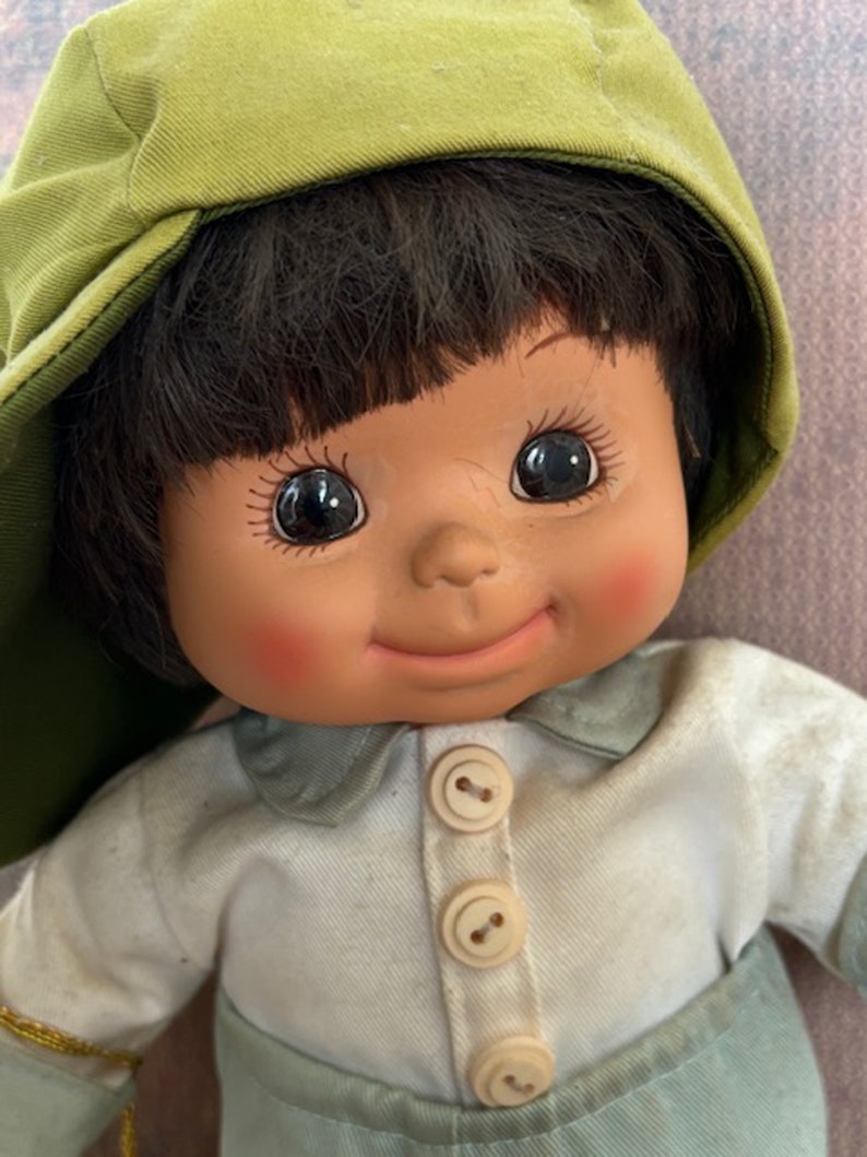 Puede incluir: Una mu&ntilde;eca sonriente con cabello casta&ntilde;o que lleva una gorra verde y una camisa azul claro con botones blancos.