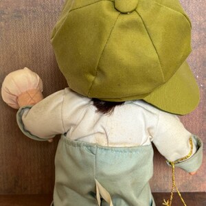 Puede incluir: Una mu&ntilde;eca de peluche con una gorra verde, una camisa blanca y pantalones azules. La mu&ntilde;eca sostiene un cono de helado blanco y marr&oacute;n.