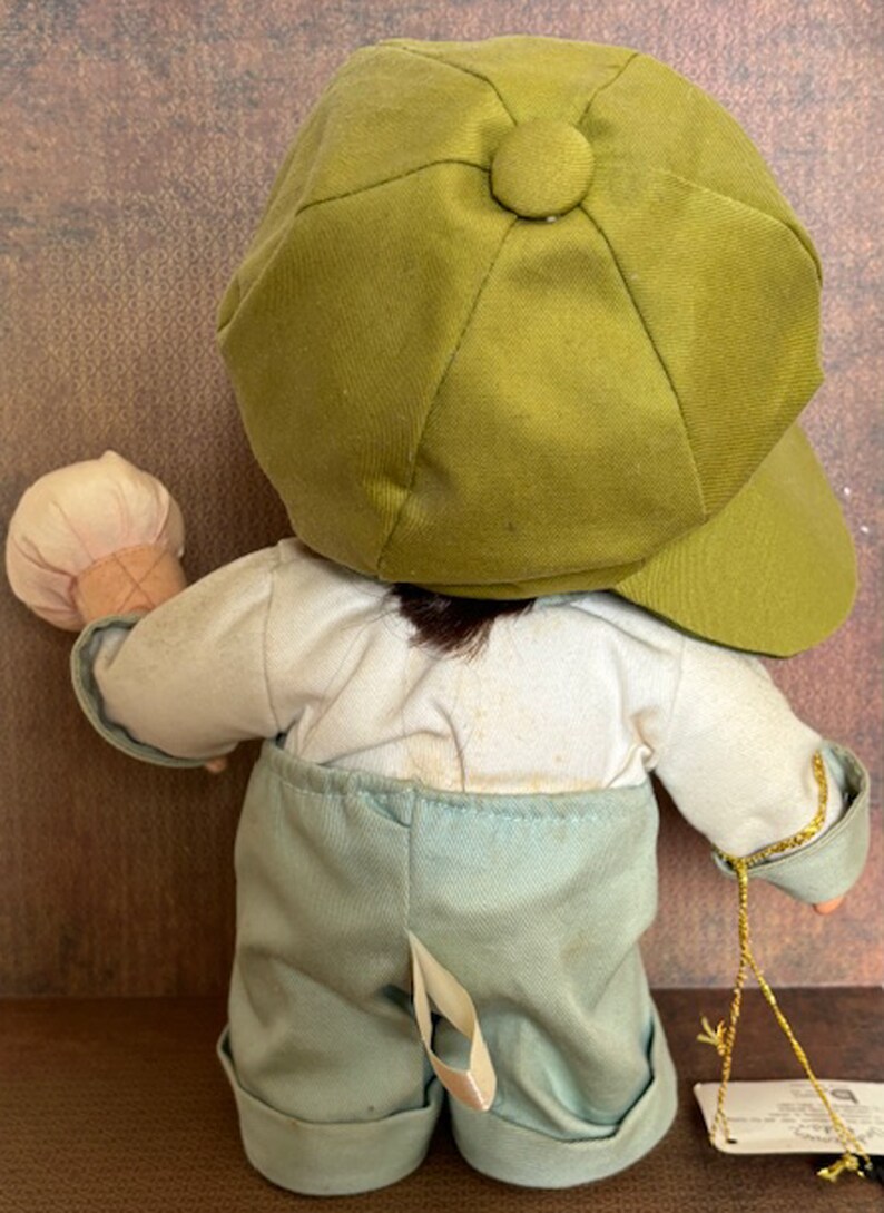 Puede incluir: Una mu&ntilde;eca de peluche con una gorra verde, una camisa blanca y pantalones azules. La mu&ntilde;eca sostiene un cono de helado blanco y marr&oacute;n.