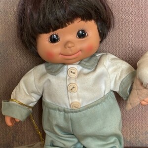 Puede incluir: Una mu&ntilde;eca de peluche con pelo casta&ntilde;o, que lleva una camisa blanca con botones y pantalones azules. La mu&ntilde;eca sostiene un cono de helado.