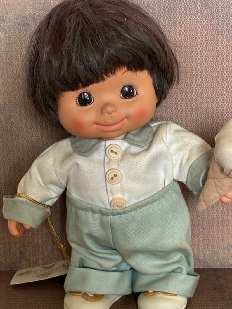 Puede incluir: Una mu&ntilde;eca de peluche con pelo casta&ntilde;o, que lleva una camisa blanca con botones y pantalones azules. La mu&ntilde;eca sostiene un cono de helado.