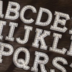 May include: Close-up of decorative alphabet letters. Each letter is white with a border of small, clear rhinestones and filled with faux pearls. The letters are arranged on a dark brown, textured surface.