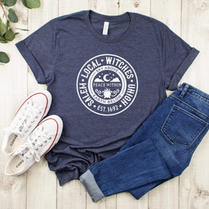 May include: A navy blue t-shirt with a white circular graphic that reads "Salem Local Witches Union" with the text "Sky Above, Peace Within, Earth Below, Est. 1692". The shirt is paired with blue jeans and white sneakers.