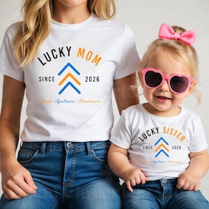 May include: Two white t-shirts with matching designs. One shirt says "LUCKY MOM" with a blue and orange arrow design and "Down Syndrome Awareness." The other shirt says "LUCKY SISTER" with the same design.