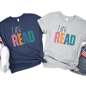 May include: Two t-shirts, one navy blue and one grey, each with the text "Let's READ" in colourful block letters. The navy shirt has a white tag at the neckline. The grey shirt has a white tag at the neckline.