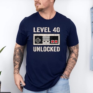 May include: Navy blue t-shirt with a white graphic of a retro video game controller and the text "LEVEL 40 UNLOCKED". The controller design features a black background with white and red details, perfect for gamers celebrating a milestone birthday.