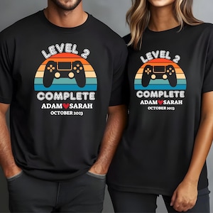 May include: Black t-shirts with a retro design featuring a game controller, a sunset, and the text "LEVEL 2 COMPLETE" with the names "Adam & Sarah" and the date "October 2023".