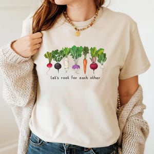 May include: Beige t-shirt featuring a colorful illustration of various root vegetables, including radishes, carrots, and beets. The text "Let's root for each other" is printed below the vegetables. The shirt is made of a soft material.