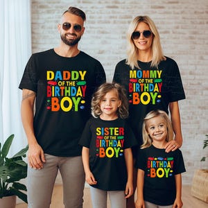 May include: Four black t-shirts with colorful text that reads "DADDY OF THE BIRTHDAY BOY", "MOMMY OF THE BIRTHDAY BOY", "SISTER OF THE BIRTHDAY BOY", and "BIRTHDAY BOY". The shirts are worn by a family of four, with the parents wearing sunglasses.