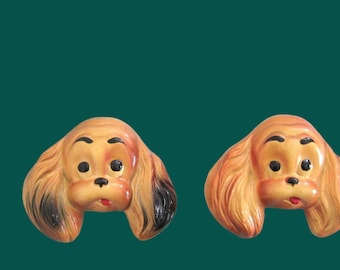 Peut inclure: Deux décorations murales en forme de tête de chien en céramique. Les chiens ont une fourrure beige, des yeux noirs et une bouche rouge. Les oreilles ont des pointes noires. Le fond est de couleur vert d'eau.