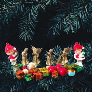 May include: A festive Christmas arrangement featuring miniature gold angel figurines, elf ornaments in red and white, and colorful wrapped gift boxes. The scene is set against a backdrop of green pine needles, creating a holiday atmosphere.