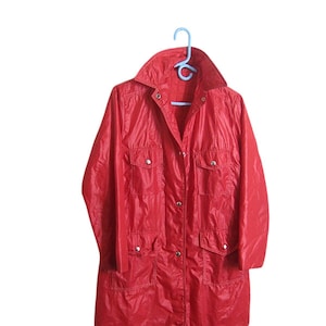May include: A bright red, lightweight, water-resistant jacket featuring a pointed collar, long sleeves, and a front button closure. The garment includes two chest pockets and two lower pockets, each secured with a silver-toned metal snap button.