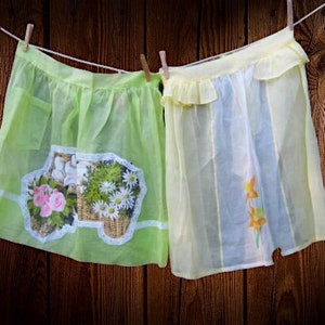 Peut inclure: Deux tabliers vintage suspendus à une corde à linge sur fond de bois. L'un est vert clair avec un motif floral, l'autre jaune pâle avec un volant et un motif de fleurs peintes. Les deux sont fixés à la corde avec des pinces à linge en bois.