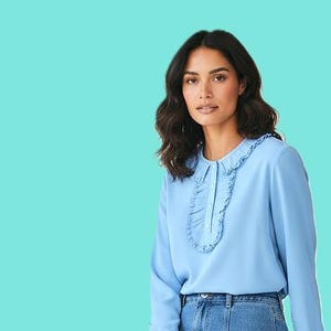 May include: A light blue blouse with a ruffled collar and cuffs. The long-sleeved top has a button-down front and is paired with a denim skirt. The model is sitting in front of a turquoise background.