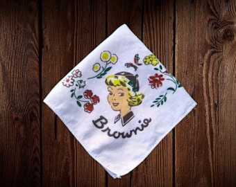 May include: White handkerchief with a vintage illustration of a woman with blonde hair and the name "Brownie" in script. The handkerchief is decorated with floral and bird designs in red, yellow, and green.