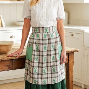 May include: A woman wearing a white blouse with lace trim and a plaid apron with green windmill designs. The apron has green pockets and is paired with a green skirt. The kitchen setting includes shelves with plates and a wooden table.