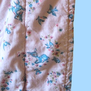 Puede incluir: Primer plano de una tela rosa con un patrón repetido de ciervos azules, pájaros, mariposas y elementos florales. La tela está acolchada con costuras verticales. El tema general es vintage y caprichoso.