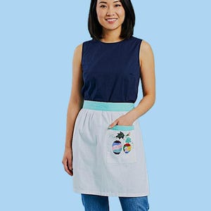 Puede incluir: Un delantal blanco con cinturilla y bolsillo azul claro. El bolsillo presenta un diseño bordado de huevos coloridos y decorados. La persona de la imagen lleva una camiseta azul marino y vaqueros azules.