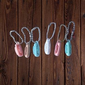 Puede incluir: Siete llaveros de teléfono rotatorio de estilo vintage en varios colores pastel, incluyendo rosa, melocotón, azul claro y blanco, colgados de cadenas plateadas sobre un fondo de madera marrón.