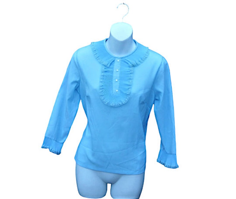 K&ouml;nnte beinhalten: Hellblaue Bluse mit R&uuml;schenkragen und -front. Die langen &Auml;rmel enden in R&uuml;schenb&uuml;ndchen. Die Bluse hat eine Reihe kleiner, wei&szlig;er Kn&ouml;pfe auf der Vorderseite und wird auf einer wei&szlig;en Schaufensterpuppe pr&auml;sentiert.