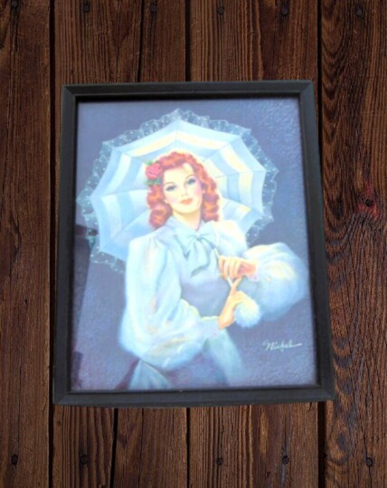 Op de afbeelding: Een ingelijste vintage print van een vrouw in een witte jurk en een witte kantparasol. Ze heeft rood haar en houdt een klein boeket bloemen vast. De print is rechtsonder getekend met "Nakaba".
