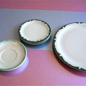 May include: A collection of white ceramic plates and saucers with a green scalloped edge. The set includes a large oval plate, two small plates, and a saucer. The plates are arranged on a pink and silver surface.