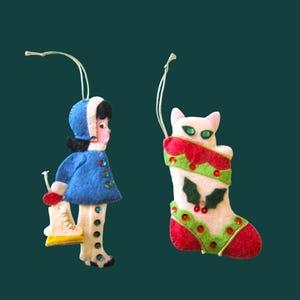 Könnte beinhalten: Zwei Filzanhänger, bestehend aus einer Figur in blauem Mantel und weißer Hose mit einem weißen Schlittschuh sowie einer weißen Katze, die aus einem rot-grünen Weihnachtsstrumpf mit Stechpalmenblättern, roten Beeren und Pailletten schaut.