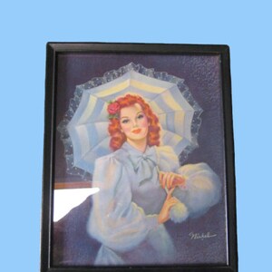 Op de afbeelding: Ingelijst vintage druk van een vrouw met rood haar die een witte jurk draagt en een witte en blauwe parasol vasthoudt. De vrouw heeft een roos in haar haar en glimlacht. De print is gesigneerd "Nickel".