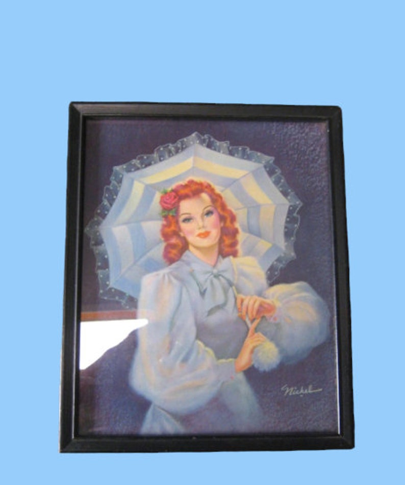 Op de afbeelding: Ingelijst vintage druk van een vrouw met rood haar die een witte jurk draagt en een witte en blauwe parasol vasthoudt. De vrouw heeft een roos in haar haar en glimlacht. De print is gesigneerd "Nickel".
