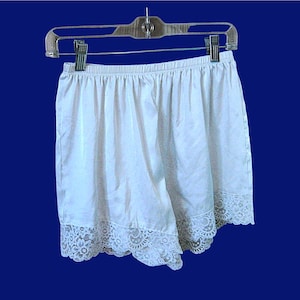 Puede incluir: Pantalones cortos de satén blanco con un ribete de encaje en el dobladillo, colgados de una percha de plástico transparente. Los pantalones cortos tienen una cintura elástica y un diseño ligeramente fruncido. El fondo es azul oscuro liso.