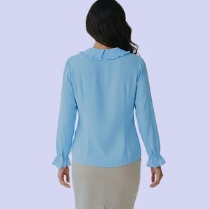 K&ouml;nnte beinhalten: Hellblaue Bluse mit langen &Auml;rmeln und R&uuml;schenkragen und -manschetten. Die Bluse ist aus einem weichen, flie&szlig;enden Stoff gefertigt und hat ein klassisches, feminines Design. Das Model tr&auml;gt einen beigen Rock.