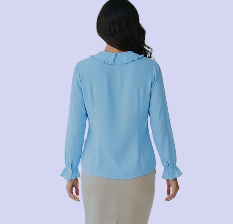 K&ouml;nnte beinhalten: Hellblaue Bluse mit langen &Auml;rmeln und R&uuml;schenkragen und -manschetten. Die Bluse ist aus einem weichen, flie&szlig;enden Stoff gefertigt und hat ein klassisches, feminines Design. Das Model tr&auml;gt einen beigen Rock.