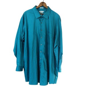 May include: A long-sleeve button-down shirt in a solid teal color. The shirt features a standard collar, a front button placket with matching teal buttons, and a single chest pocket on the left side. It is displayed on a wooden clothes hanger.