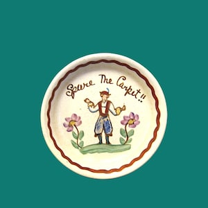 Peut inclure: Une coupelle en céramique ronde à bord brun festonné, ornée d'une illustration peinte à la main représentant une personne en tenue traditionnelle sur un monticule vert entre deux fleurs violettes, avec le texte "Spare The Carpet!!".
