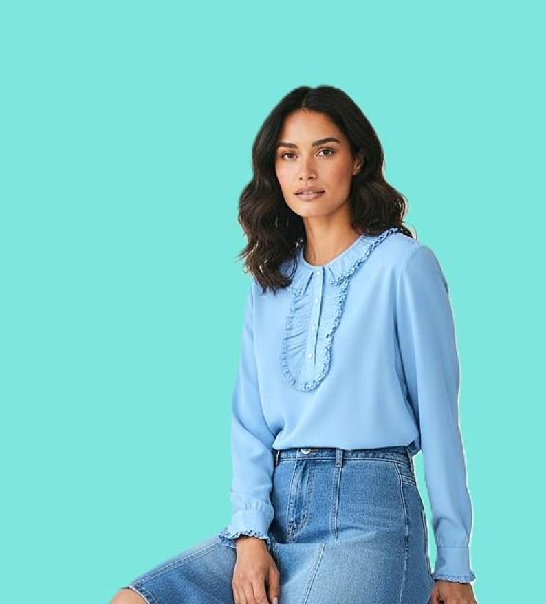 May include: A light blue blouse with a ruffled collar and cuffs. The long-sleeved top has a button-down front and is paired with a denim skirt. The model is sitting in front of a turquoise background.