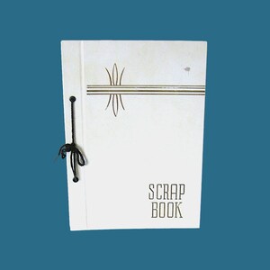 Puede incluir: Un álbum de recortes blanco con un cierre de cuerda negra. La portada presenta un diseño dorado de líneas y un motivo floral, con las palabras "SCRAP BOOK" impresas en una fuente decorativa. El álbum de recortes está sobre un fondo verde azulado.