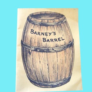 Può includere: Una stampa su carta color marrone chiaro con un'illustrazione a inchiostro dettagliata di un barile di legno con cerchi in metallo scuro. Il testo BARNEY'S BARREL è stampato in nero al centro del barile, che presenta un piccolo foro circolare.