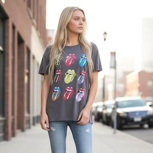 May include: A gray t-shirt featuring multiple colorful tongue and lip designs. The shirt has a classic crew neck and short sleeves. The design includes red, blue, yellow, and purple lips. The model is wearing light blue jeans.