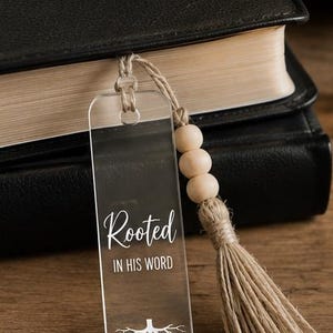Peut inclure: Un marque-page rectangulaire en acrylique transparent avec le texte imprimé en blanc Rooted IN HIS WORD, un graphique blanc de racines d'arbre et le texte COLOSSIANS 2:7 en bas. Il comporte un pompon en ficelle de jute et trois perles en bois clair.