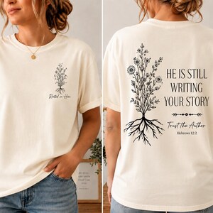 May include: An off-white short-sleeve t-shirt featuring a small black floral graphic and the text Rooted in Him on the front left chest. The back displays a large black floral graphic with the text HE IS STILL WRITING YOUR STORY Trust the Author Hebrews 12:2.