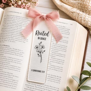 Peut inclure: Un marque-page rectangulaire translucide avec le texte noir "Rooted IN GRACE" et "2 CORINTHIANS 12:9" au-dessus et en dessous d'une illustration au trait noir d'une petite tige fleurie. Un ruban rose poudré effiloché est noué au sommet.