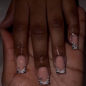 Może przedstawiać: Zbliżenie dłoni z kwadratowymi paznokciami. Paznokcie mają jasnoróżową bazę z białym wzorem french manicure. Końcówki zdobione są czarnymi paskami zebry.