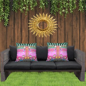 May include: A black three-seater outdoor sofa with two pink and white patterned throw pillows. The sofa is set against a wooden fence with greenery hanging from the top. A gold sunburst mirror hangs on the wall behind the sofa. Two topiary trees are on either side of the sofa.