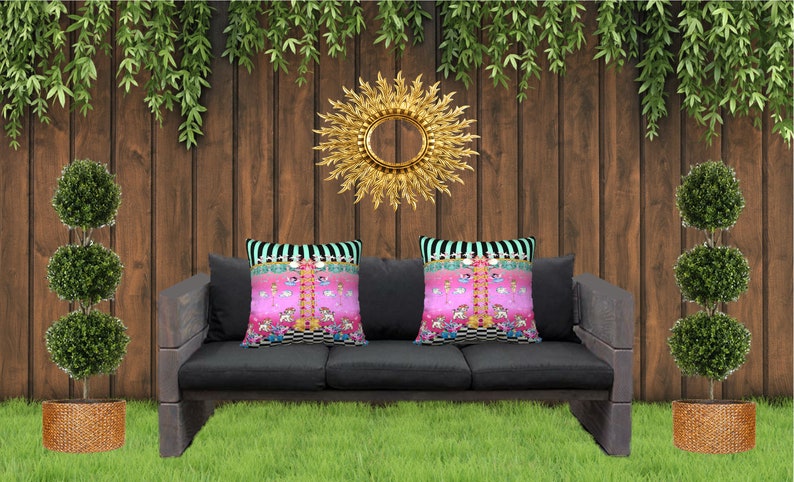 May include: A black three-seater outdoor sofa with two pink and white patterned throw pillows. The sofa is set against a wooden fence with greenery hanging from the top. A gold sunburst mirror hangs on the wall behind the sofa. Two topiary trees are on either side of the sofa.