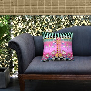 May include: A blue velvet sofa with a pink and black patterned throw pillow featuring a whimsical design of dancing figures and animals. The sofa is in a room with a wooden floor and a green leafy plant in a pot.
