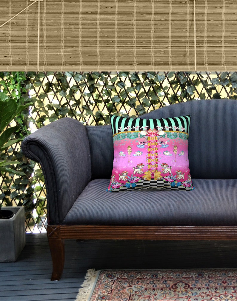 May include: A blue velvet sofa with a pink and black patterned throw pillow featuring a whimsical design of dancing figures and animals. The sofa is in a room with a wooden floor and a green leafy plant in a pot.