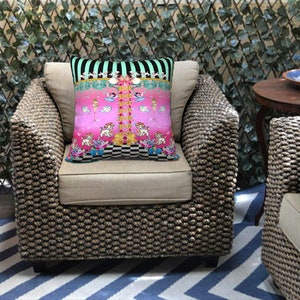 May include: A pink and green patterned throw pillow with a whimsical design featuring a carousel with animals and a checkered floor. The pillow is on a wicker chair with a striped rug.